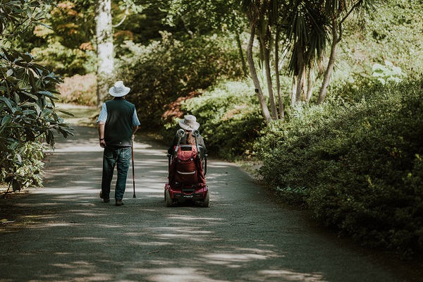 Un couple de personnes se promenant dans un endroit verdoyant, la femme est dans un fauteuil roulant et l'homme marche avec une canne